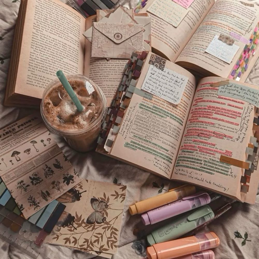 Books with colorful annotations and tabs, an iced coffee, pastel highlighters, and decorative paper sheets on a floral bedspread.