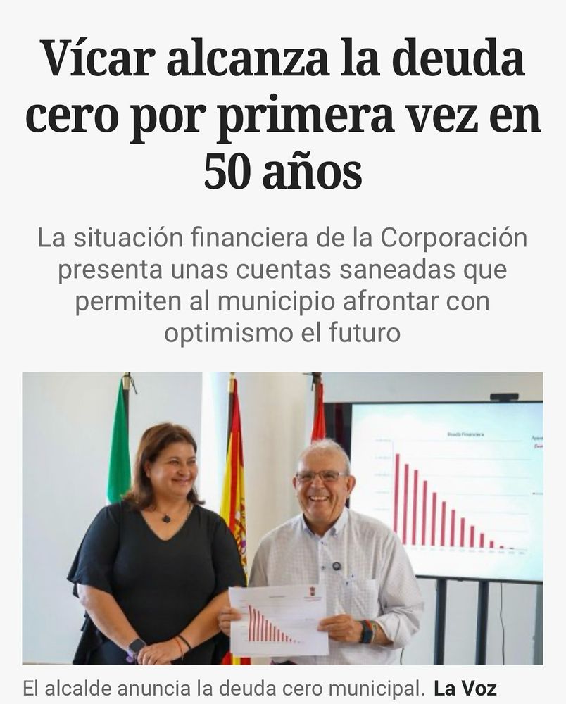 Fotografía de dos personas sonrientes, una mujer y un hombre, en un ambiente de oficina. El hombre sostiene un papel con una gráfica de barras descendente, mientras que detrás de ellos hay varias banderas y una pantalla que muestra una gráfica similar. El texto informa que el municipio de Vícar ha alcanzado la deuda cero por primera vez en 50 años, destacando la buena situación financiera y el optimismo para el futuro.