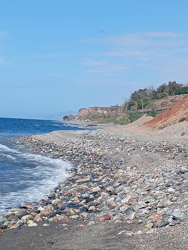 Playa de piedras y arena, con olas suaves rompiendo en la orilla. A la derecha, hay un acantilado con vegetación y algunos árboles. Al fondo, se observa una carretera que sigue la línea de la costa y algunas construcciones sobre el acantilado. El cielo está despejado con algunas nubes dispersas y el mar es de un azul intenso. La imagen transmite tranquilidad y naturaleza costera.