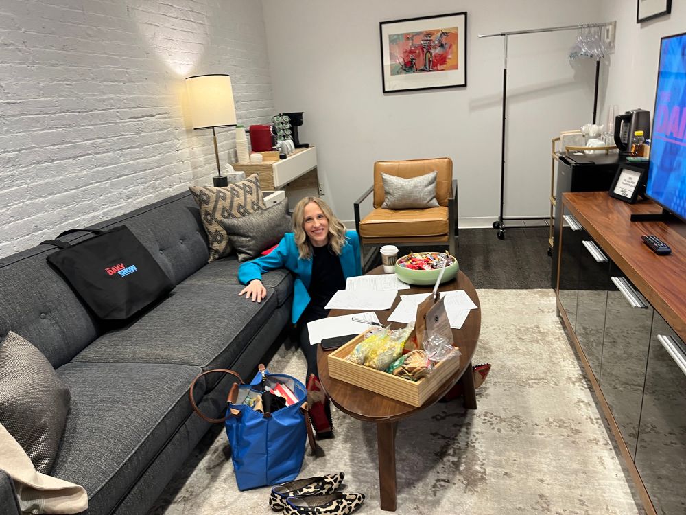 Author, Umich law professor, and cohost of the Strict Scrutiny podcast Leah Litman smiling and sitting on the floor in a turquoise blazer surrounded by pieces of paper and a giant bowl of snacks, her arm leaning on a grey couch, backstage before her appearance on The Daily Show.