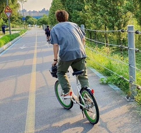 Namjoon riding a bike on the bike path of a park. He has his back turned to the camera. He has brown hair and is wearing a gray shirt and dark green pants.