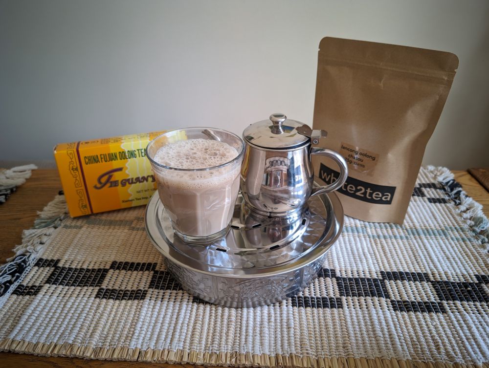 a yellow box of tieguanyin next a stainless steel tray and teapot, with a glass of frothy light brown milk tea, next to a brown paper bag