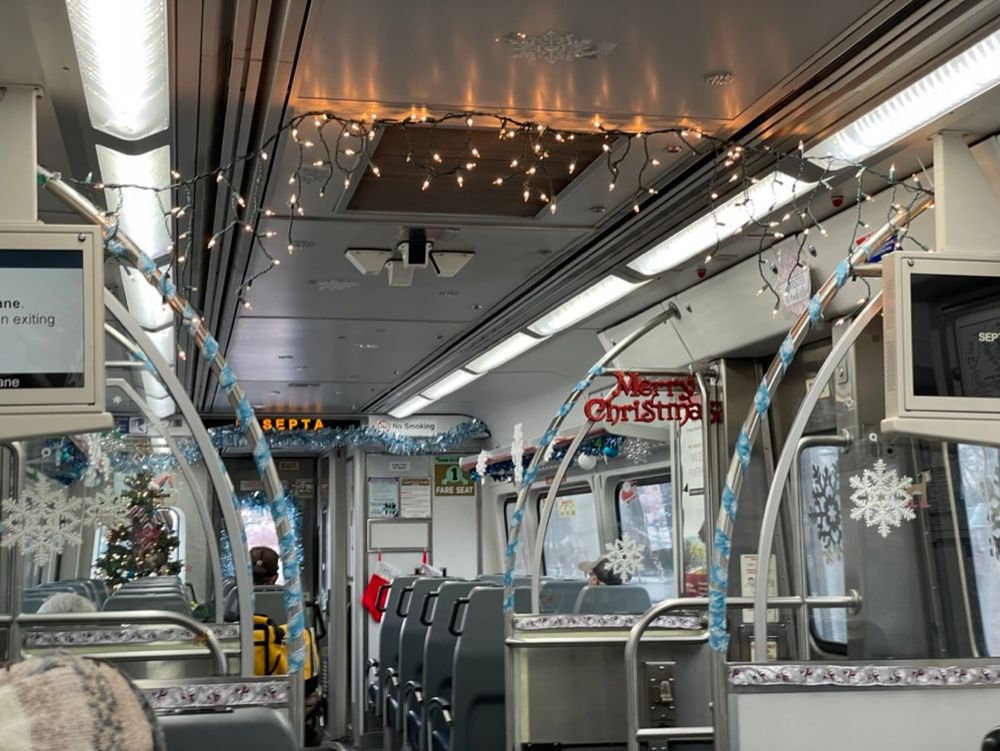 The inside of a SEPTA commuter rail car. The inside has been decorated with string lights, snowflake decals, merry christmas signs, and garlands. It's quite excessive, but very charming!