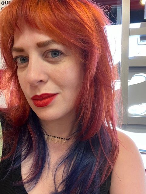 A white woman with red and purple hair, red lipstick, and blue eyes. While she has dark circles, she does not look half as bad as the corrupt dude in the photo below her.