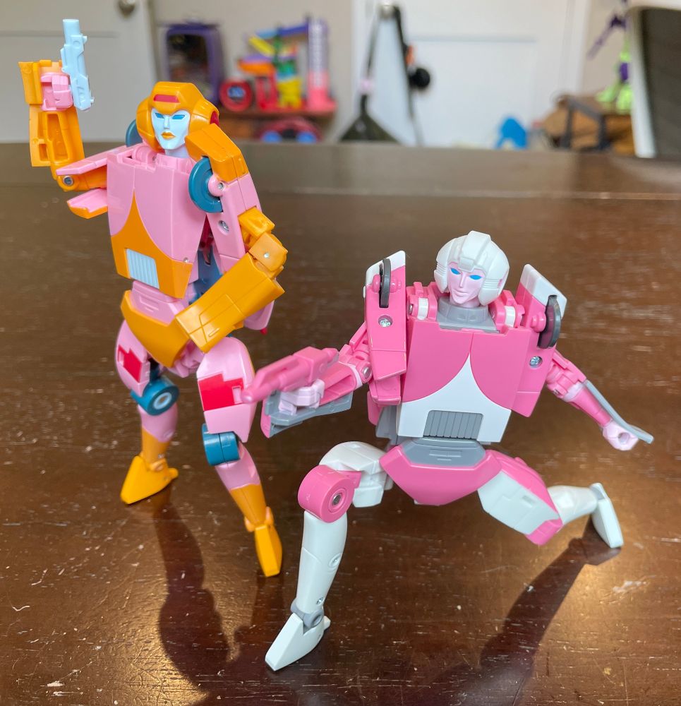 The two Missing Link Arcee figures is more elaborate poses. The pink/orange Arcee is looking off to the side while holding up a laser gun in her right hand. Her left hand is resting on her hip.

The pink/white Arcee is crouched down with her right leg bent and her left leg stretched out behind her. She is taking aim at something off camera with the laser gun in her right hand.