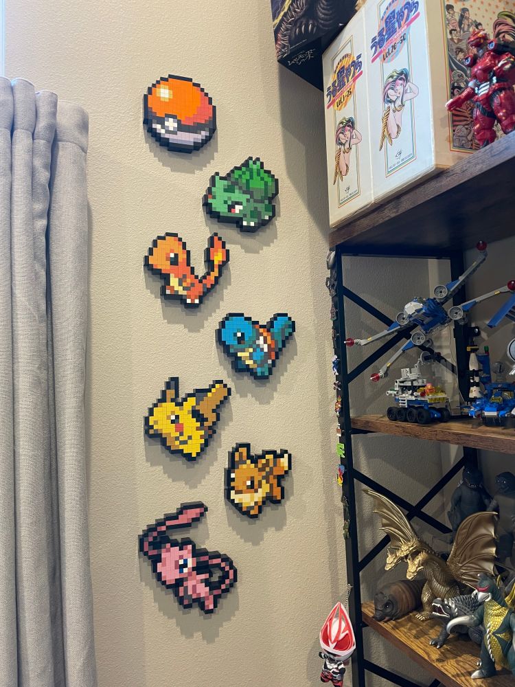 A collection of Mattel's MEGA Pokémon Pixel Art building sets hung vertically on a wall. There's a Poké Ball, Bulbasaur, Charmander, Squirtle, Pikachu, Eevee, and Mew.