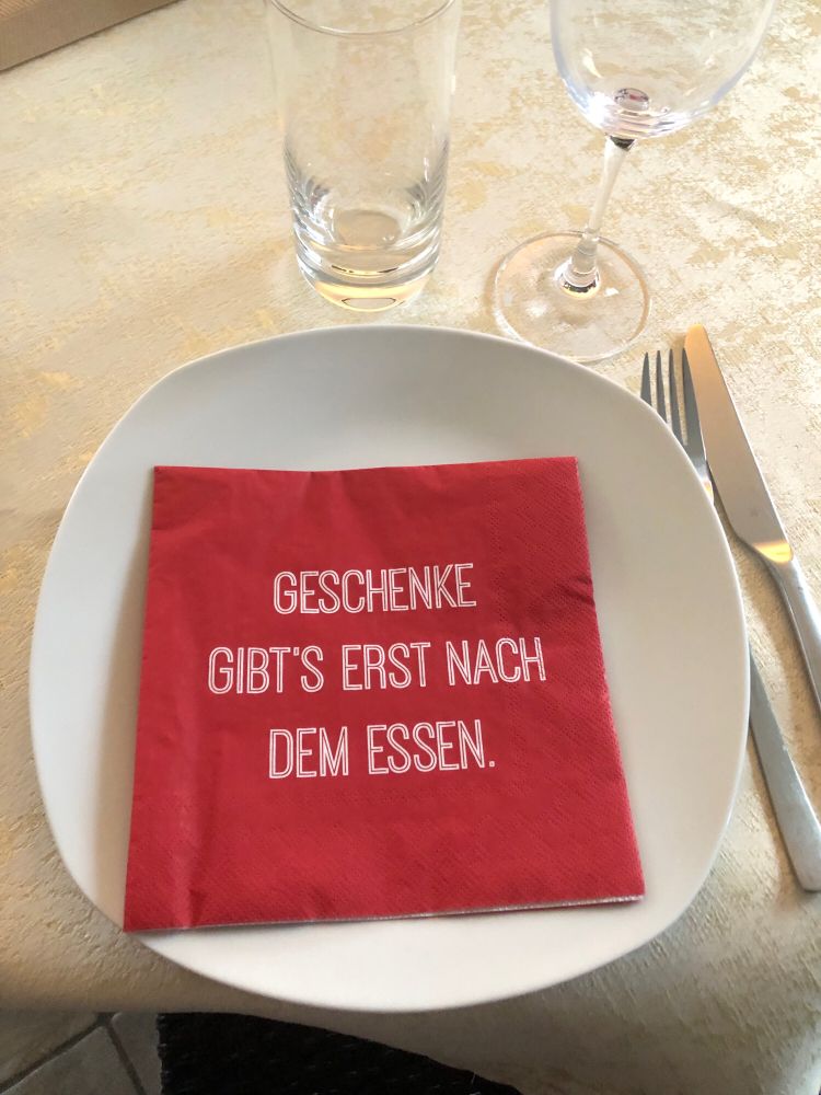 gedeckter Tisch, auf dem Teller liegt eine Serviette mit folgendem Text: Geschenke gibt‘s erst nach dem Essen“