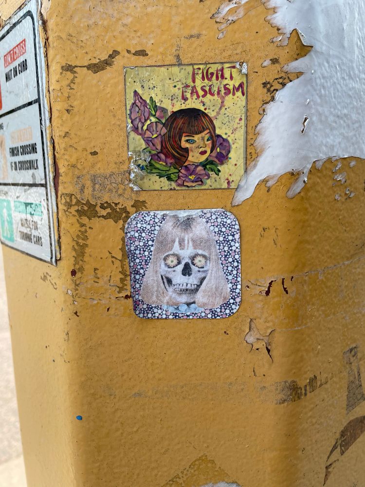 Two stickers on a slightly rusty and chipped traffic light pole. The one on top is in a classic tattoo style with a blue eyed person with brown hair in a 1920s style bob cut surrounded by purple lillies with the words “fight fascism”. The sticker below it is a skull with daisies in the eye sockets and a blond bob wig 