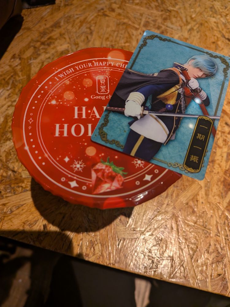 赤いフタに「I WISH YOUR HAPPY CHR...」「Gong cha」「HAPPY HOLI...」などの文字とイチゴのイラストが描かれた容器と、青髪のキャラクター「一期一振」のカードが木製テーブルの上に置かれている。