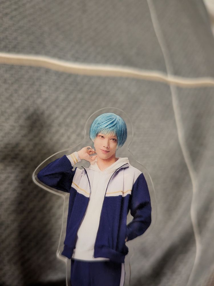 一期一振のアクリルスタンド。青い髪のキャラクターが白と紺のジャージを着て、片手を首に添えて微笑んでいる。背景はグレーの布地。