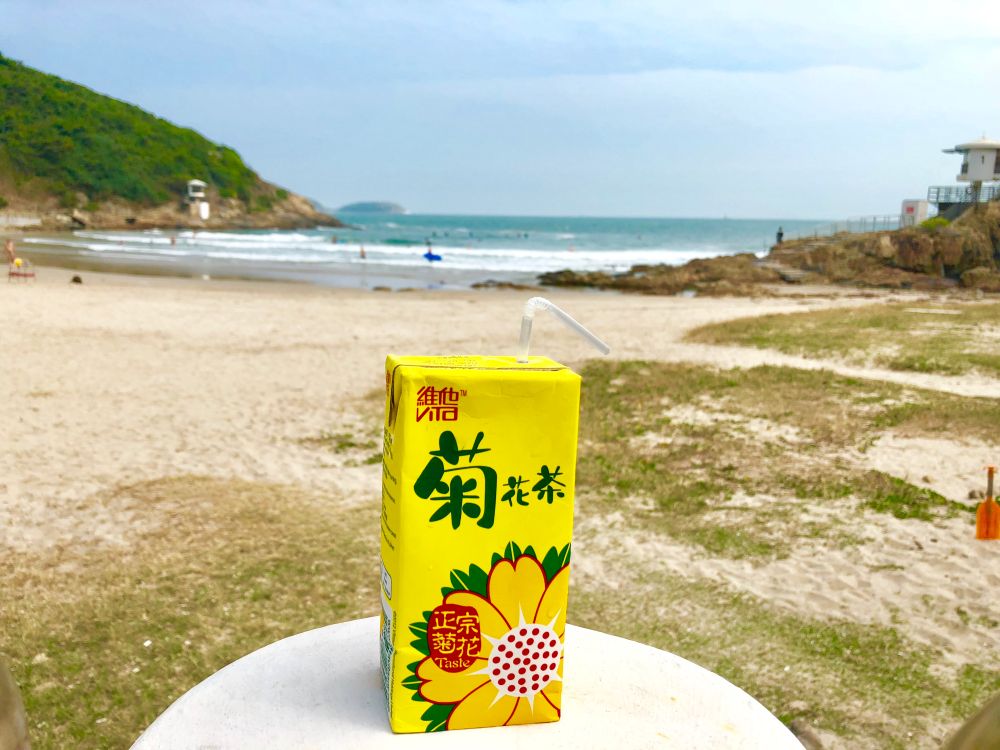 A picture of a sunny Hong Kong beach in the background with a yellow juice box on a round table in the foreground. The juice box has a yellow flower on the front and a flexible straw.