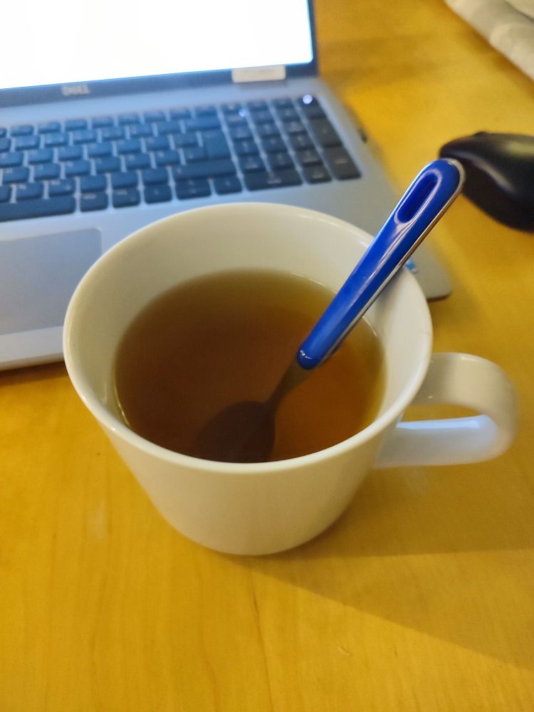Auf einem Tisch. Im Vordergrund steht eine weiße große Tasse mit einem Löffel. In der Tasse ist Tee. Dahinter steht ein Notebook. 