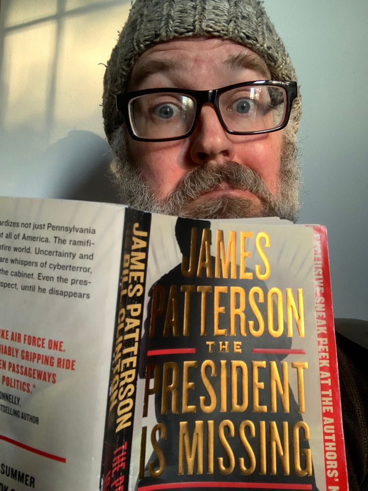 A guy with a smirk holding a book with “The President is Missing” in sparkly gold lettering. 