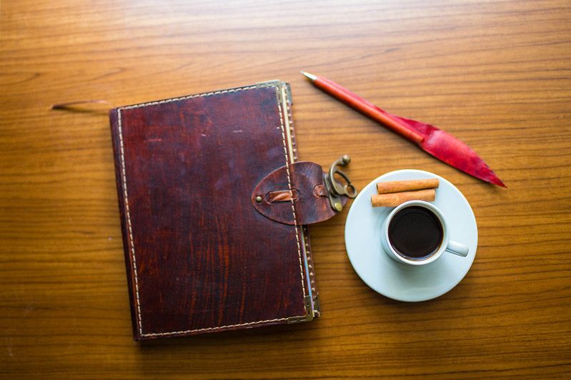 A leather-bound notebook and a pen on a table, with a cup of coffee and some finger biscuits