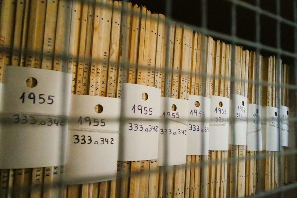 A row of old documents stored vertically on shelves behind a metal grid. Each file has a white tag with handwritten text reading '1955' and '333a342'.
