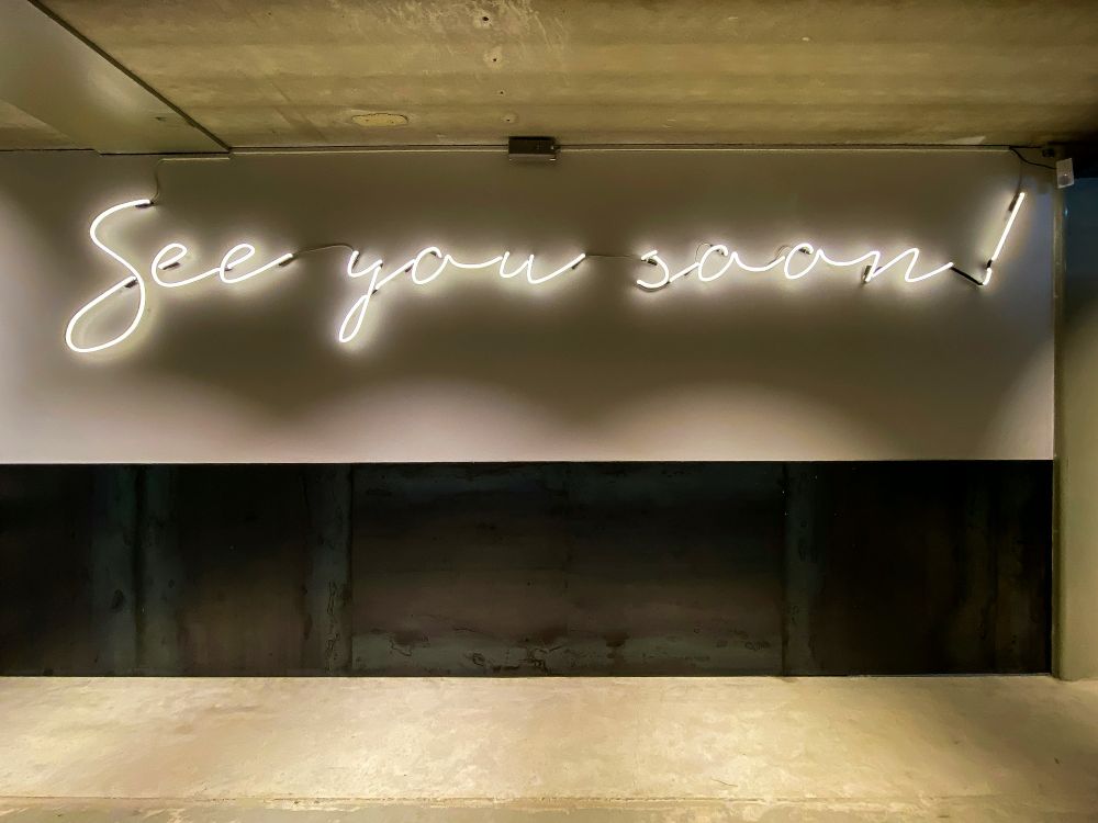 A neon sign on a wall reads 'See you soon!' in cursive handwriting. The sign is illuminated with white light, and the background consists of a concrete wall and ceiling with an industrial appearance.