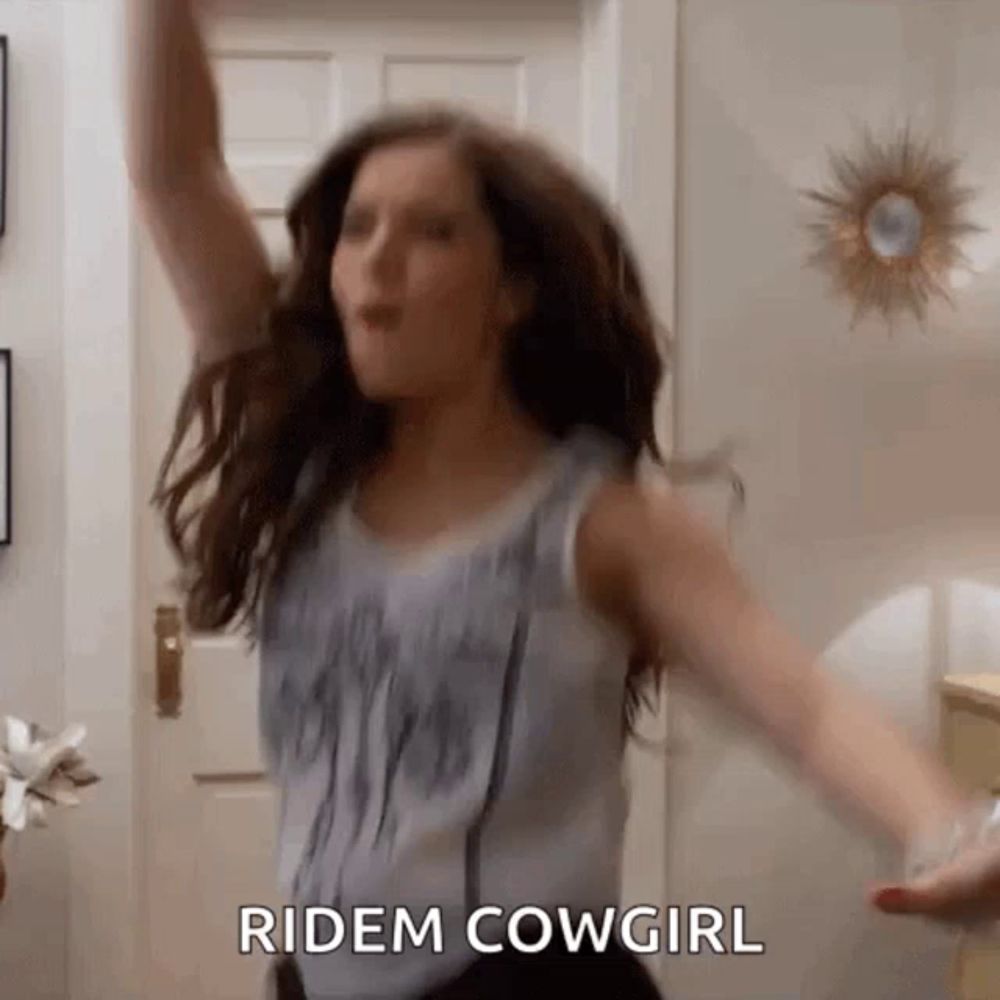 a woman is dancing in a living room with her arms outstretched and the words ridem cowgirl written on the screen .