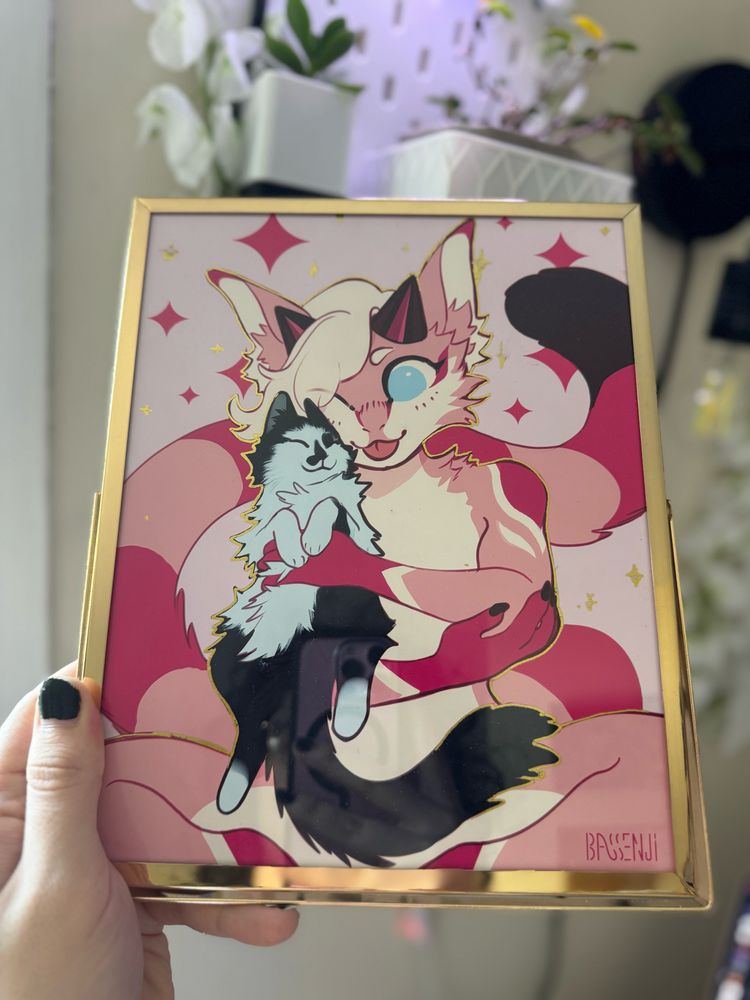 A print of my fursona Paris holding my IRL cat skunk. It's in a gold frame with gold, reflective outlines in the piece. She is holding him up to her face and nuzzling him. Paris has her tongue out and is nuzzling him, while Skunk has a very content look on his face. There are pink and gold sparkles in the background.