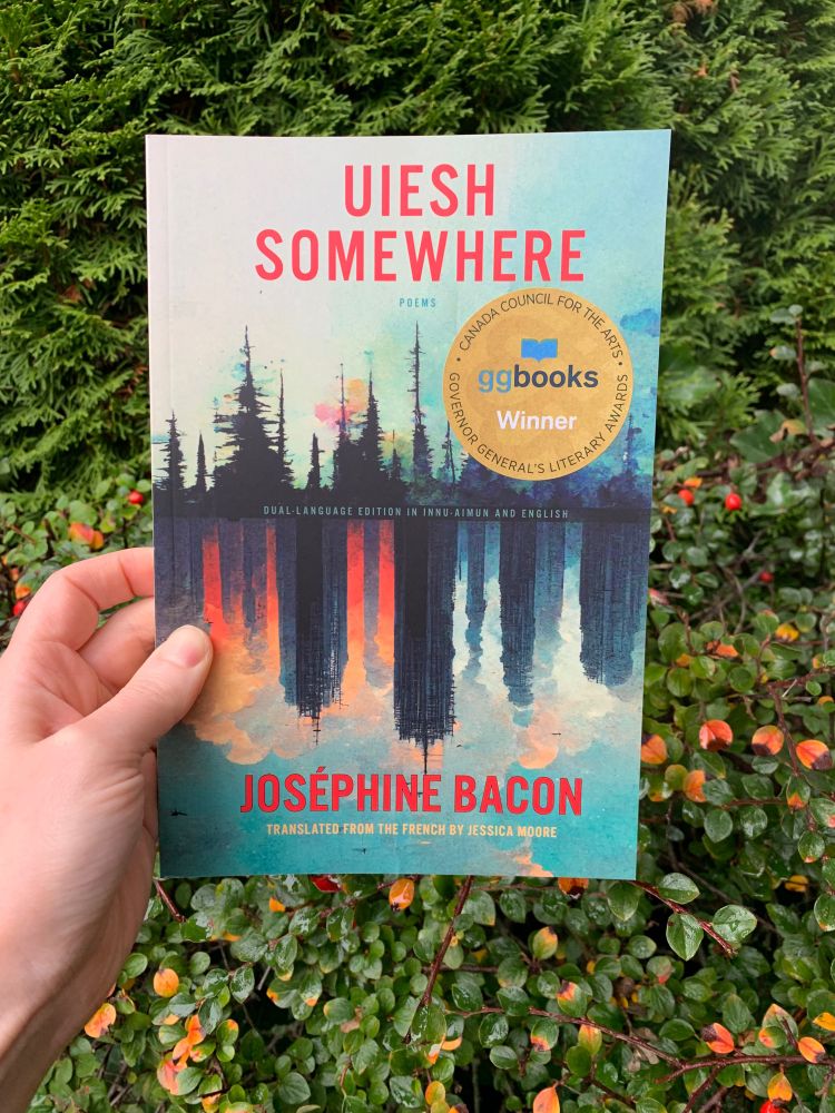 Against a backdrop of cedar, a hand holds up Uiesh / Somewhere by Joséphine Bacon, translated by Jessica Moore. A big sticker is above the mid line to the right declaring it to be a GGBooks Winner.

The cover is of a painted treescape reflecting into water, but the reflection is a city. Across the top it reads "Uiesh / Somewhere. Poems." Across the middle it reads Dual-Language Edition in Innu-aimun and English. Across the bottom it reads "Joséphine Bacon. Translated from the French by Jessica Moore."