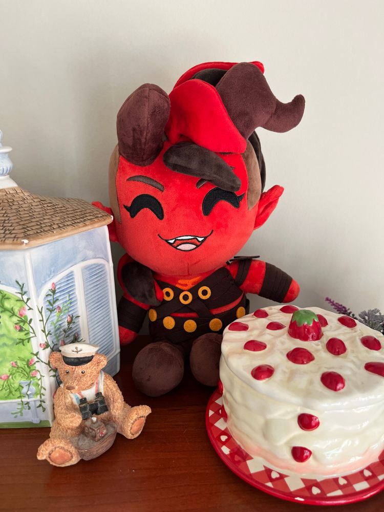 cute plush of karlach from baldur’s gate 3. karlach is a tiefling with red skin, red and black hair, and horns (one of which is broken). she is sitting on a shelf next to a small figurine of a teddy bear dressed as a sailor, a candle jar that looks like a strawberry and whipped cream cake, and another ceramic jar that looks like a gazebo.