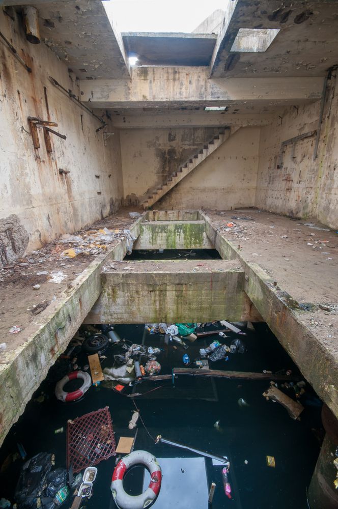 concrete steps lead down to a concrete 'bunker like' structure with open floors to the sky & sea. Flotsam of discarded junk floats in the sea below the open floor space.  
