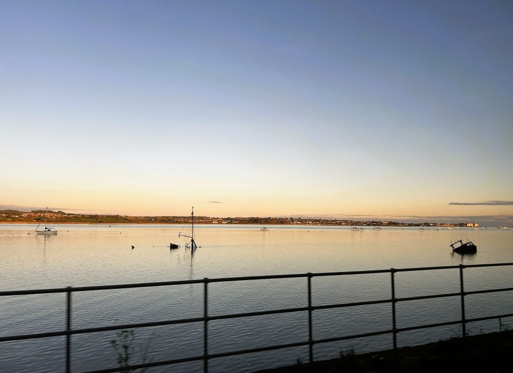 Looking east at dusk over a calm estuary. The sun is still reaching the eastern bank, making the houses of Exmouth and the hills of Woodbury Common glow orange. In the water some boats are anchored but two - a small yacht and a small motor boat - are halfsunken. Their wrecks are silhouetted by the reflected dusk light in the water.