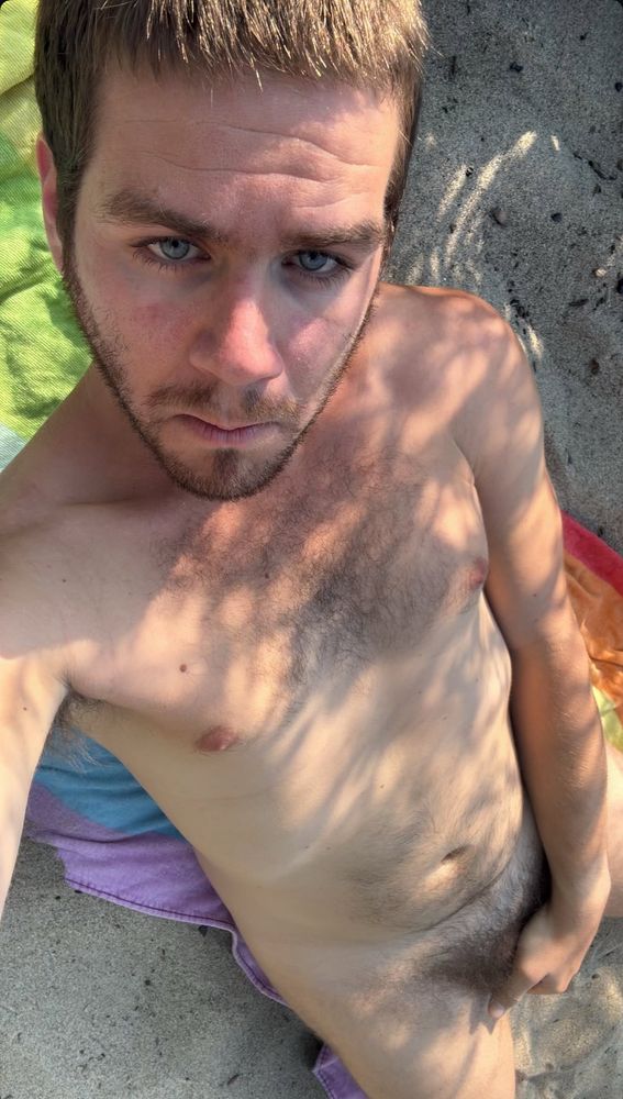 A man lying on the beach naked on a rainbow towel covering his private area 
