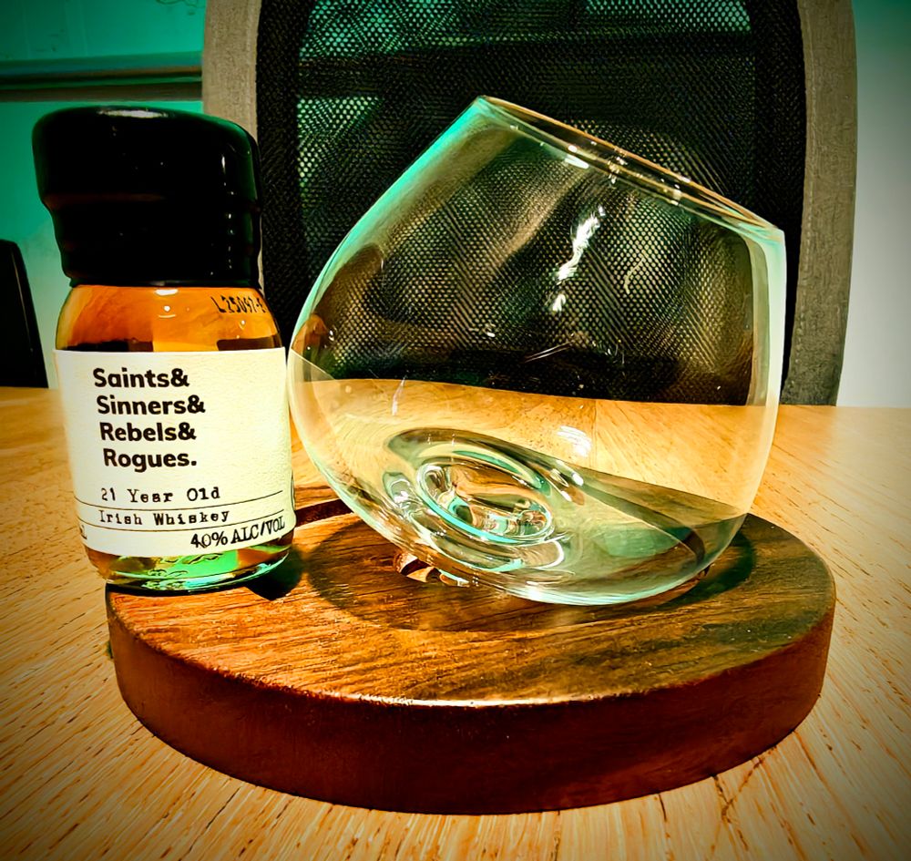 A photo of a dram of whiskey next to a round bottom glass. The whiskey is 21 year old Irish whiskey and called Saints& Sinners& Rebels& Rogues. 