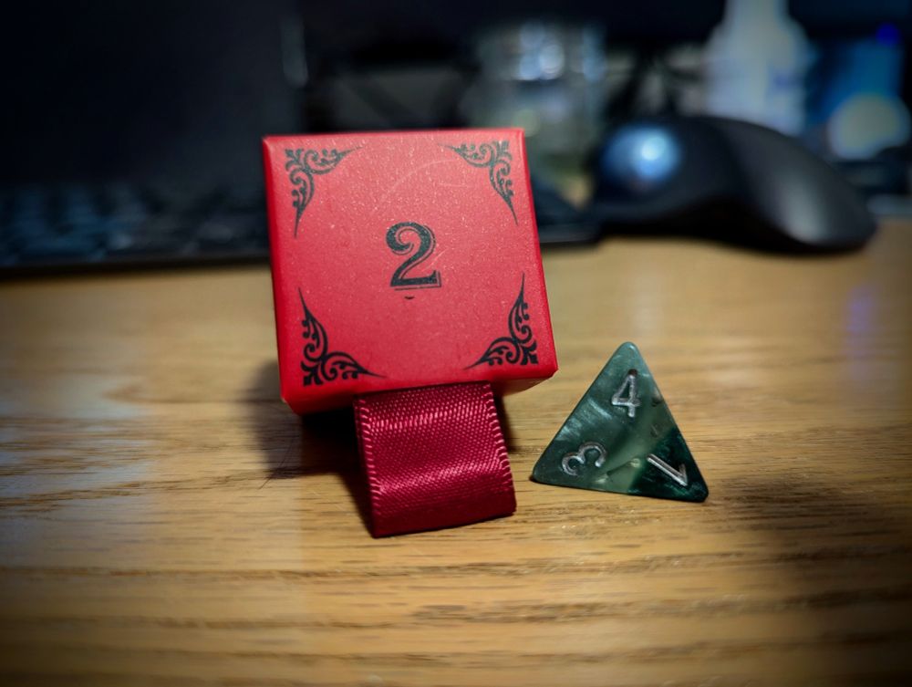 A red box with the number 2 printed in the middle. The edges of the box have a black decal. Beside the box is a green D4 dice. It's very pretty.