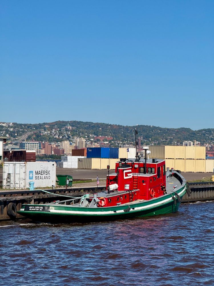 A green and red tugboat tied up along a dock stacked with shipping containers in the Duluth harbor.