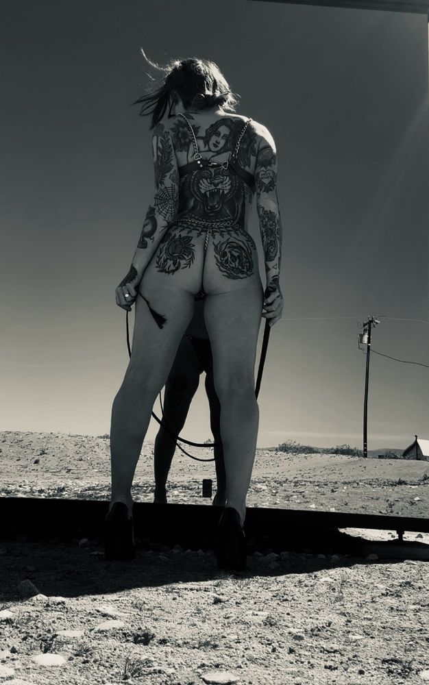 B&W image. A heavily tattooed femme stands in high heels on a desert gravel landscape. She is in front of a missed wall. The viewer can see only her bare back, covered in traditional tattoos, and a reflection of a whip, in the mirror reflection. 