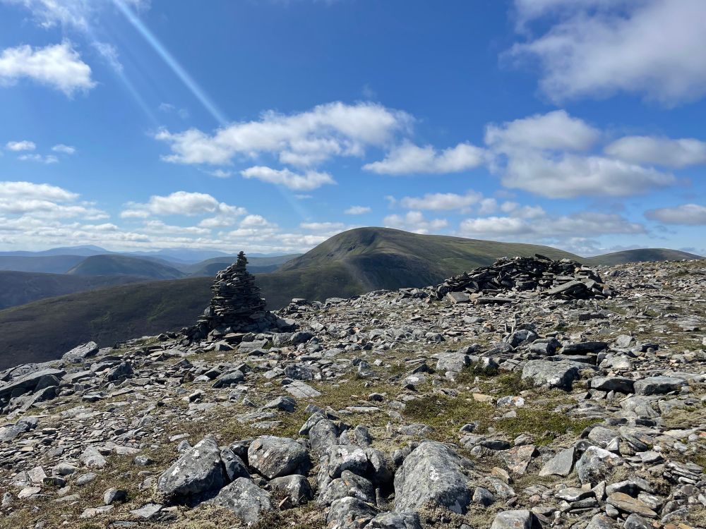 Small cairn near the summit of a Munro. Other mountains can be seen in the distance. The sky is blue with some light scattered clouds 