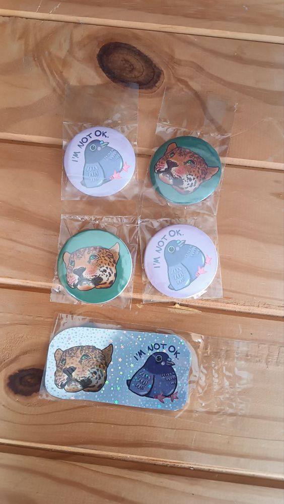 A picture showing two designs printed on pins, holo pins, and holo vinyl.
The first one is a cartoon-y pigeon in purple, sitting, that say 'I'm not ok' over her, the background is lilac.
The second one is a portrait of a yaguareté, with green background.