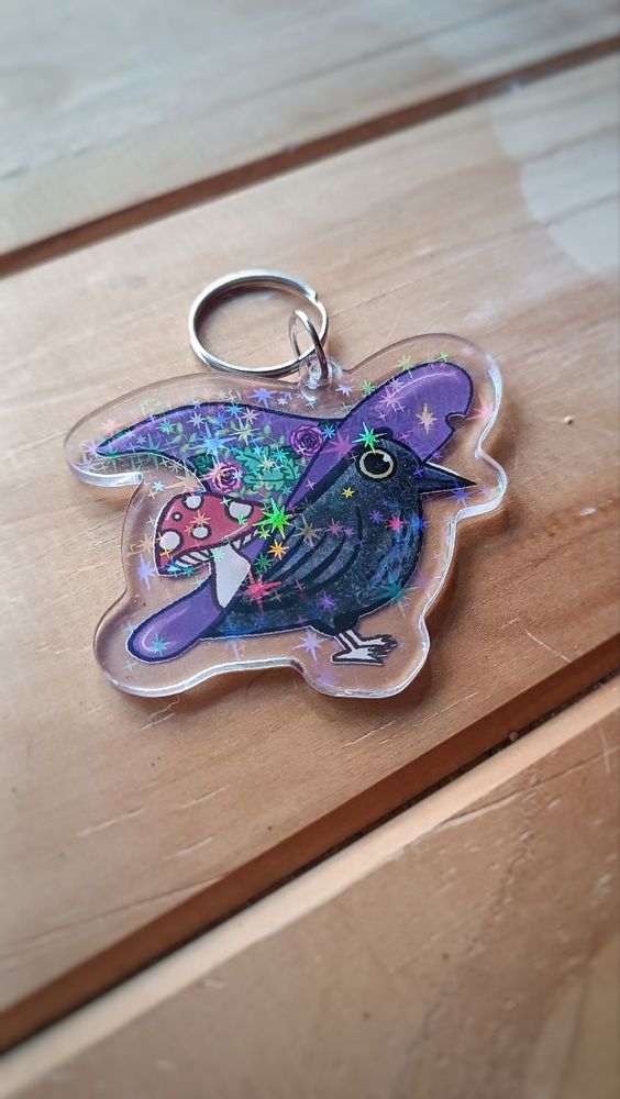 A Holo acrylic keychain depicting a raven with a huge purple witch hat. The hat has flowers, leaves, and a red mushroom.