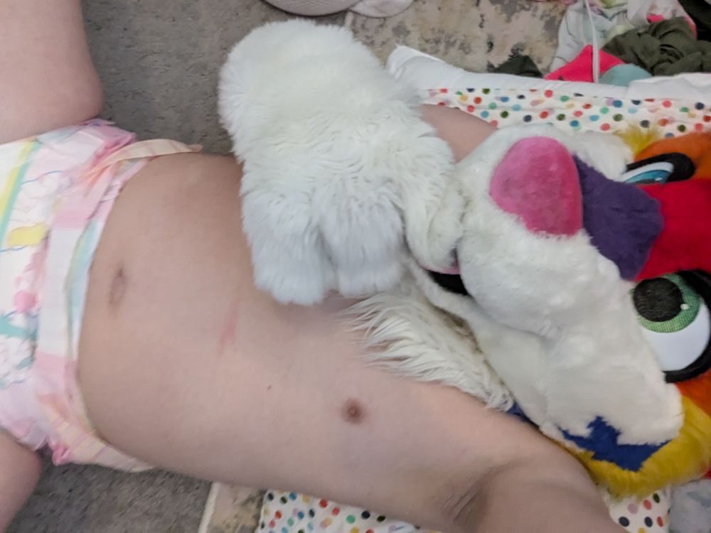 A half naked rainbow furred husky puppy looking up at the camera with a pink diaper while she nibbles on her paws cutely