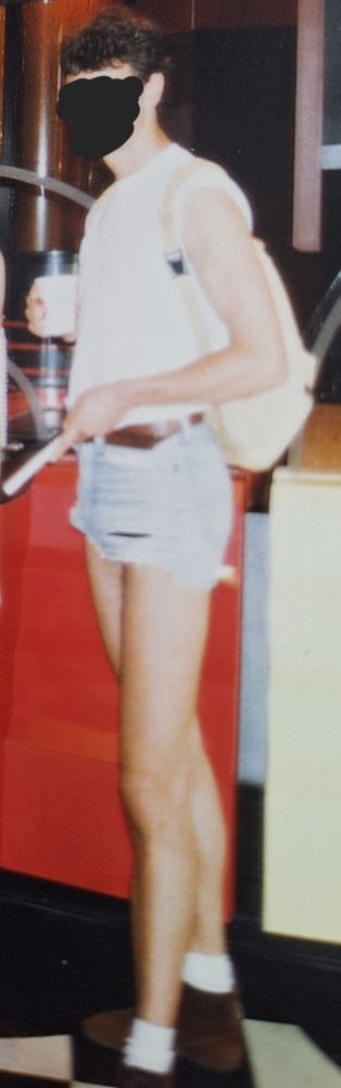 old photograph of a man white tshirt with the sleeves rolled up, a cream colored backpack, rly short light denim shorts with a brown belt, white socks and brown shoes. he is holding a travel mug in one hand and a brochure in the other