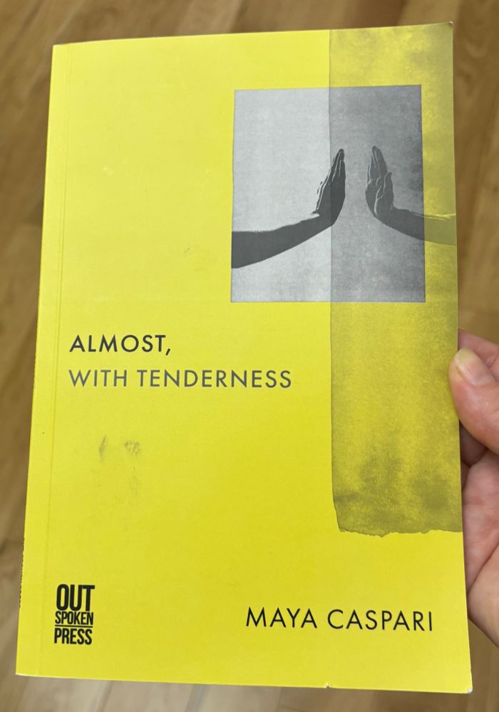 A hand holds the yellow book cover of Almost, With Tenderness by Maya Caspari from Outspoken Press. It is literally a well-thumbed book with a smudged black finger print on the front