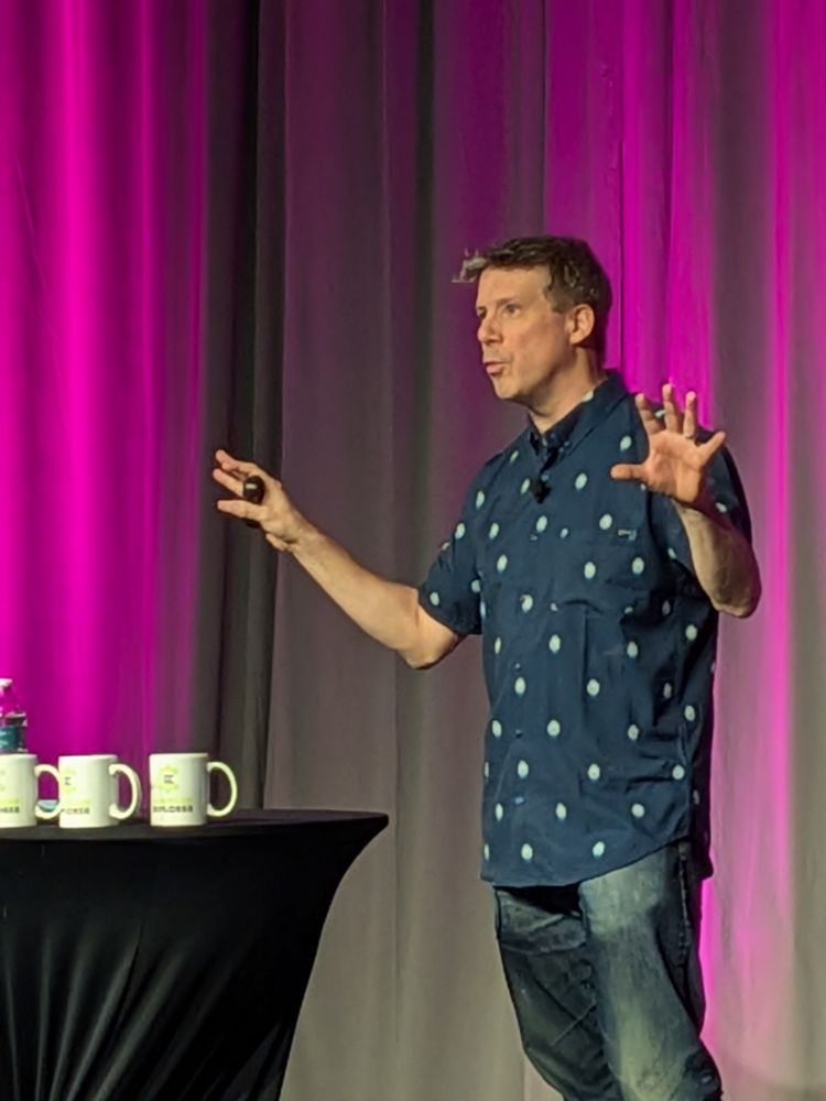 Me on stage. There's a purple light behind me, a table with compiler explorer mugs on. I'm standing mid sentence with my arms outreached in a kind of "magic trick" hocus pocus stance