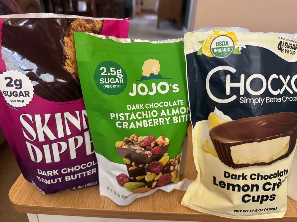 Three large bags of candy lined up in a row. Skinny Dipped Dark Chocolate Peanut Butter Cups, JoJo’s Dark Chocolate Pistachio Almond Cranberry Bites, and Chocxo Dark Chocolate Lemon Crème Cups.