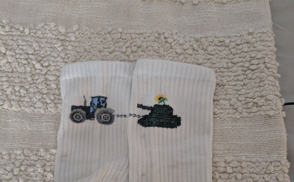A pair of white socks with a picture of a tractor towing a tank. The tank has a sunflower in it. 