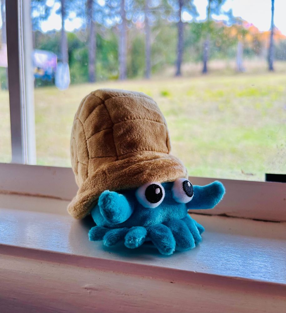 An Omanyte pokemon plush toy, sitting on a white ledge by the window. The body of Omanyte is bright blue and has many little tentacles on the bottom & 2 main arm tentacles at either side. It has 2 big googly eyes under the shell. The shell is brown and in a swirl shape. Omanyte is based on a nautilus, but looks a lot more adorable in my opinion