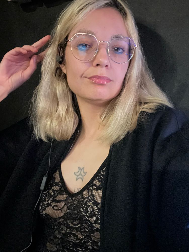 Pessoa platinada com um óculos de armação dourada, moletom de zíper aberto preto com um Body de renda preta