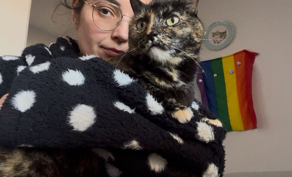 Euzinhe de Calmão preto com bolinhas brancas com uma gata belíssima de pêlo de escaminha no colo - ao fundo uma bandeira LGBT