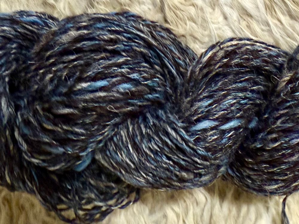 Skein of hand spun yarn on fluffy white sheepskin background. Yarn is blue and dark brown tweed, with specs of white 