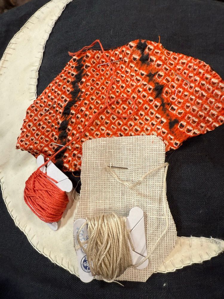 Sewing project in progress. White crescent moon on black background, with a rough cutout of an orange, black, and white mushroom cap and a tan checked mushroom stalk on top. Each mushroom part has a card of perfectly matching embroidery thread on top of it
