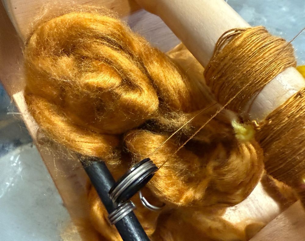 Flier of an e-spinner with very thin gold silk in the process of being spun. Unspun fiber is piled behind for emphasis 