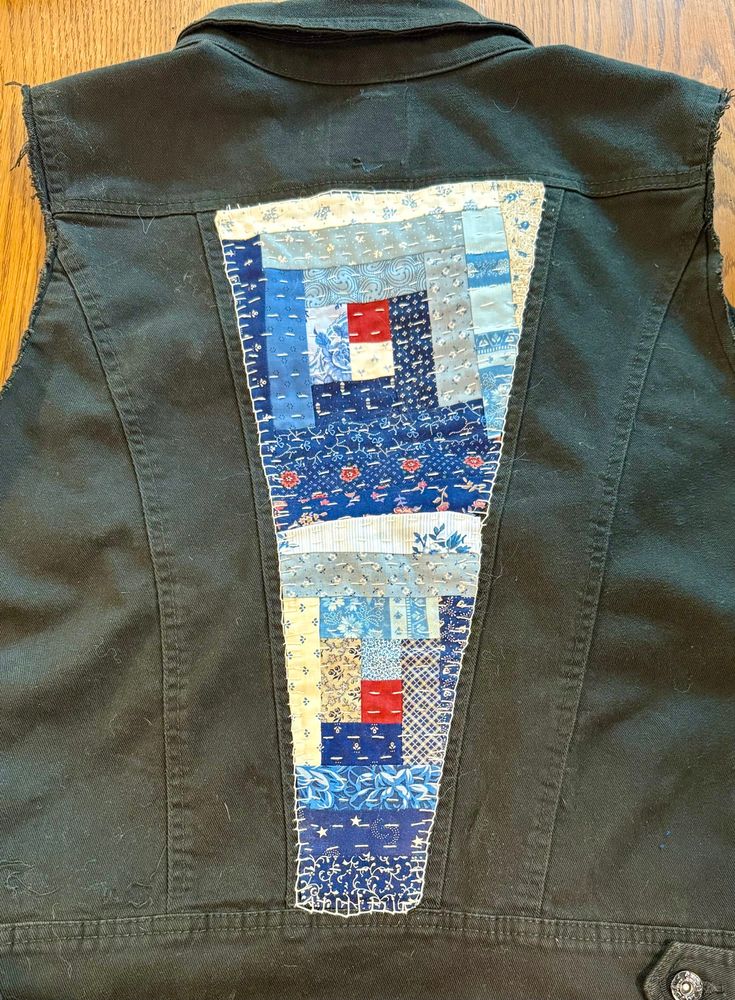 Black denim vest with quilt top back patch. Quilt top is in shades of blue in the traditional log cabin pattern, with a red center 