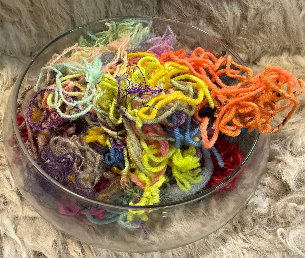 Glass bowl of multicolored yarn and roving scraps sitting on fluffy white sheepskin