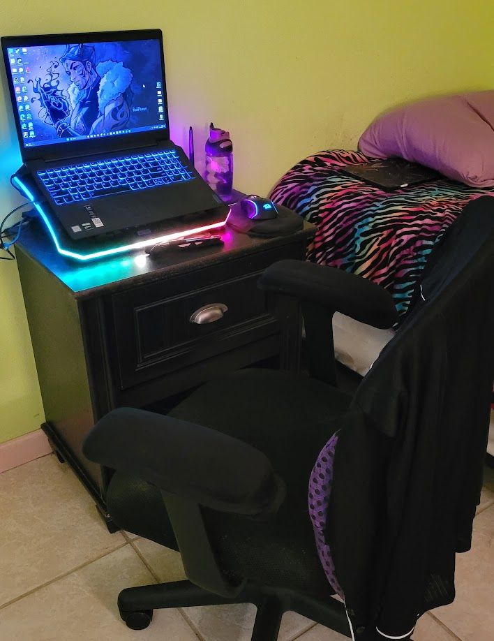 A photo of a nightstand, an office chair with a blouse on top, a laptop with a laptop cooler showing rainbow lights, an LED wired mouse, a purple water bottle and a cintiq pen next to a bed with a rainbow comforter, part of a pillow and an ipad on top.