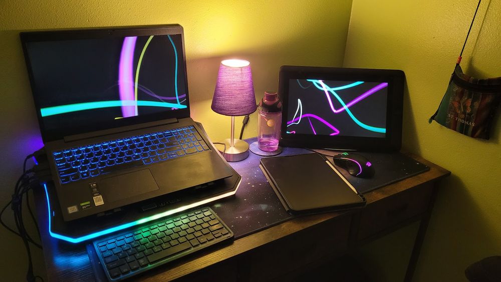 A photo of a 40" table with a laptop + laptop cooler, a cintiq 13hd, a purple lamp, an almost empty purple water bottle, an ipad + apple pencil, the same mouse from the first photo and a bluetooth keyboard on top of a purple galaxy deskmat with a st thomas wallet purse sticking from the top far right corner.